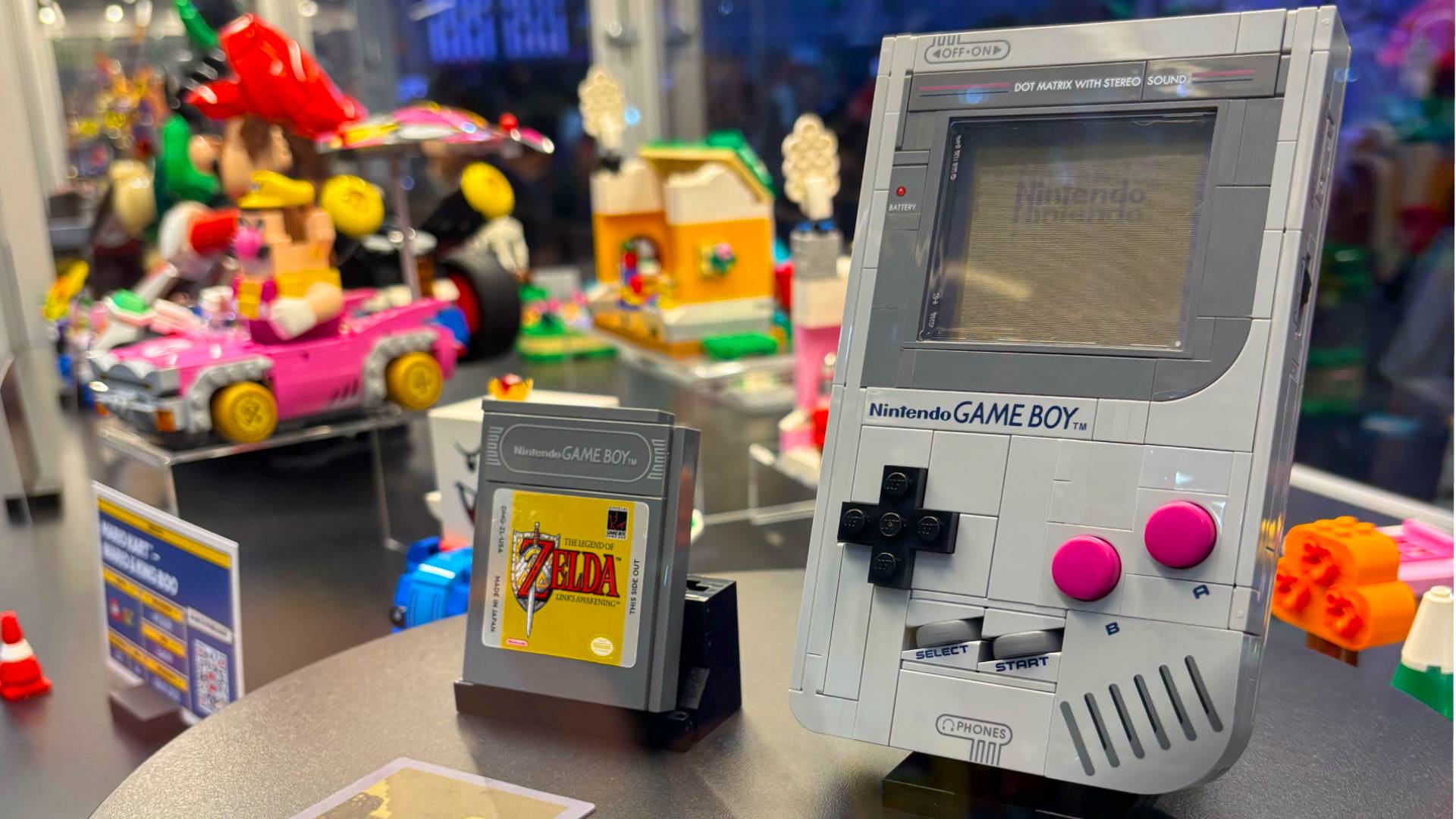 Angled photo of Lego Game Boy sitting on black table next to Zelda: Links Awakening cartridge with Mario Kart models in backdrop.