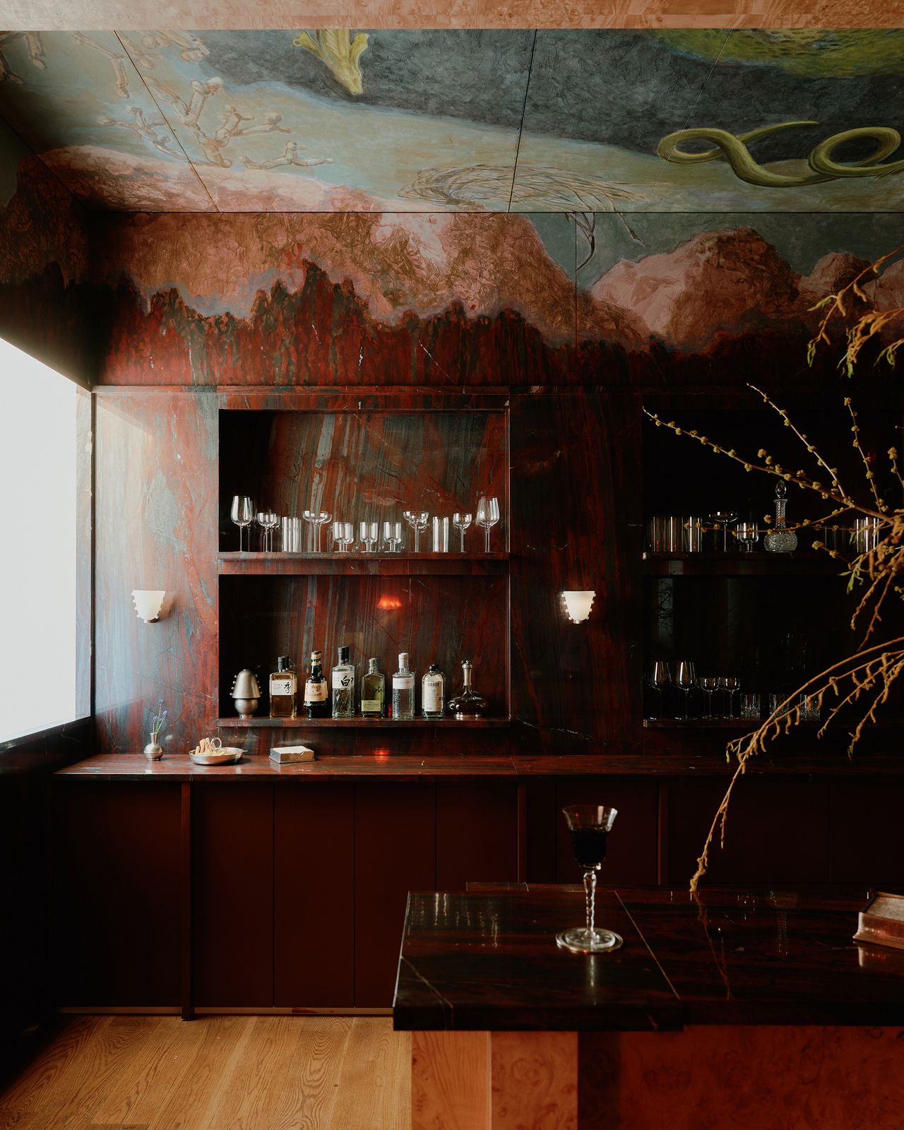 An oxblood-tinted bar counter features a whimsical ceiling mural evoking natural elements, along with sophisticated surfaces and wood tones.