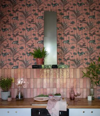 White kitchen cabinets, wood countertop, pink tile backsplash, patterned wallpaper above and extractor