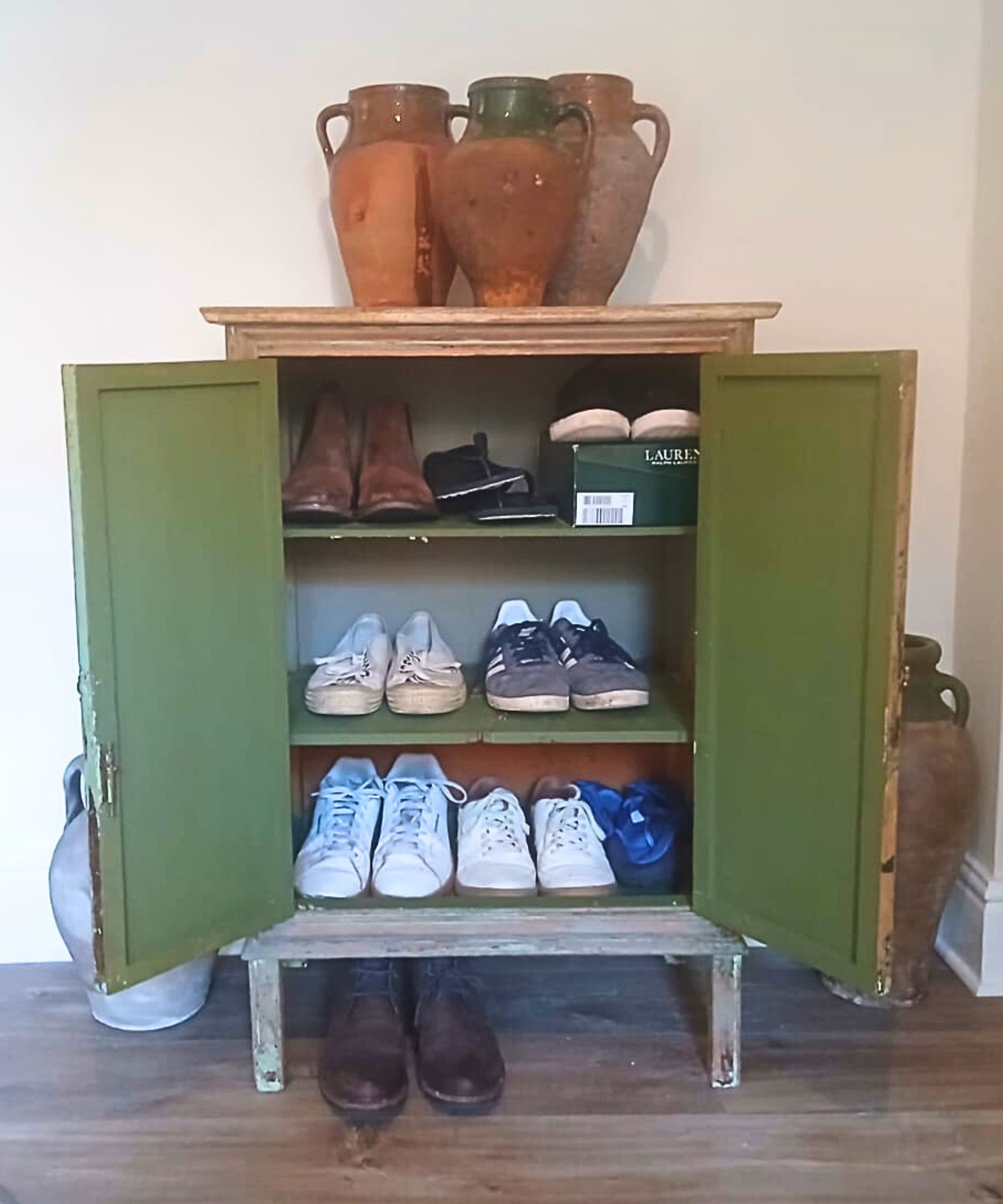 An antique green painted wooden cabinet with doors open to reveal three internal shelves neatly storing multiple pairs of shoes and trainers, with a further pair of boots tucked underneath. Three rustic terracotta amphora vessels are displayed on top, against a plain cream wall.