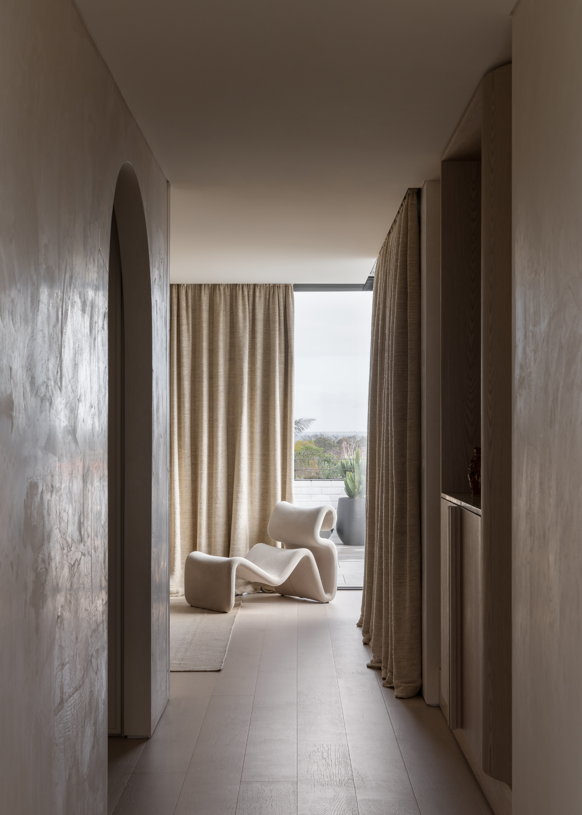 A beige passageway leading to a bright living corner with a structured chair