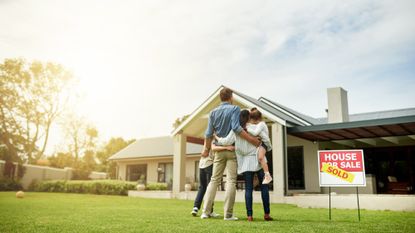 A family of four standing in the front yard of a home next to a "house for sale / sold" sign