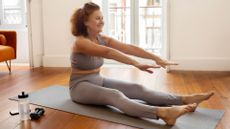 woman sat on an exercise mat on wooden flooring wearing leggings and crop top, legs outstretched and arms up at shoulder height reaching toward her feet.