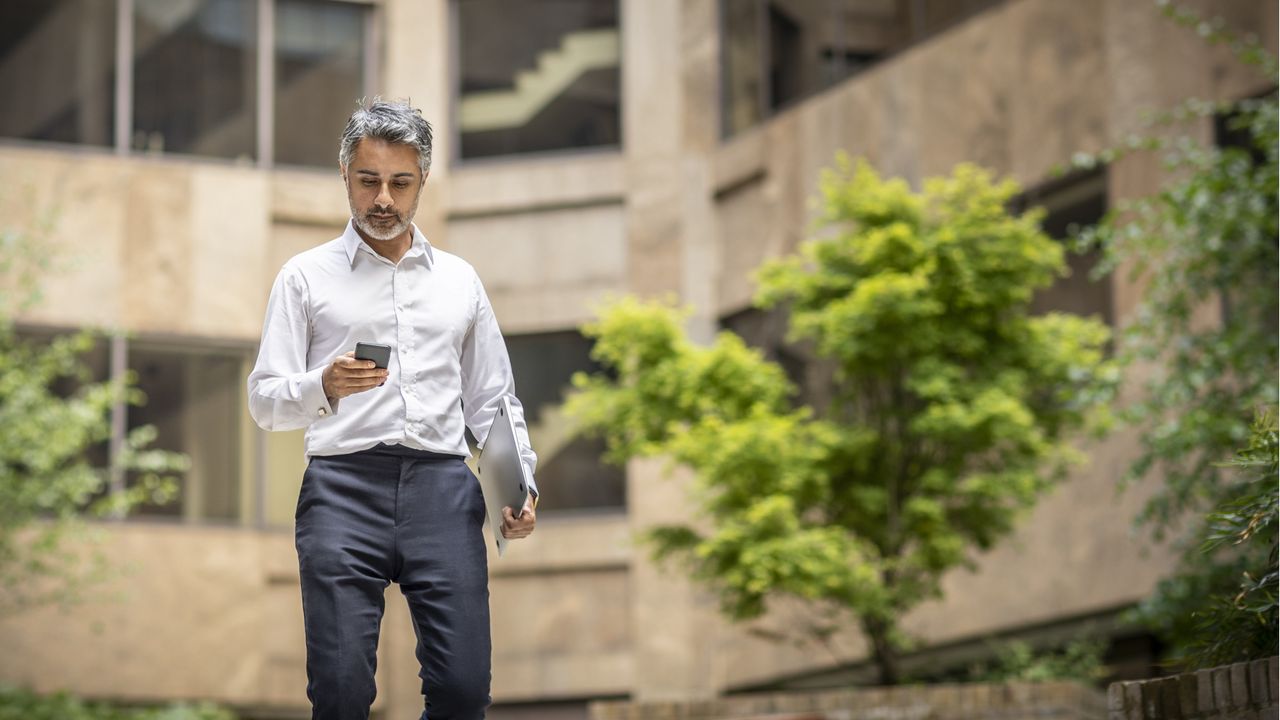 A businessman carrying a closed laptop walks outside a building while looking at his phone.