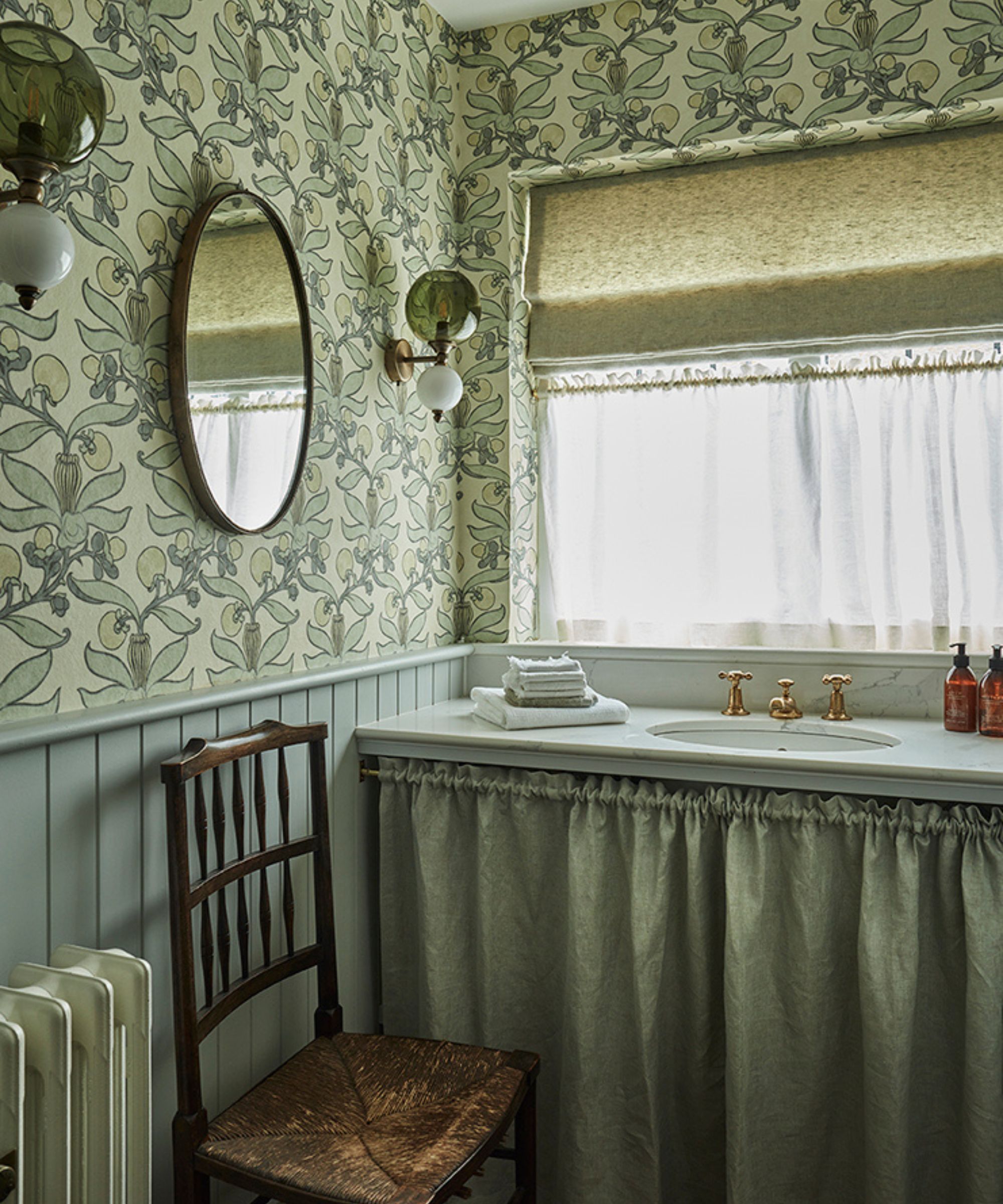 a soft blue green powder room with floral wallpaper, paneling and a skirted sink unit with a vintage chair