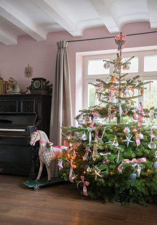 A living room with pink wall decor, black piano, rocking horse and real Christmas tree decorated with pink ribbon and star decor