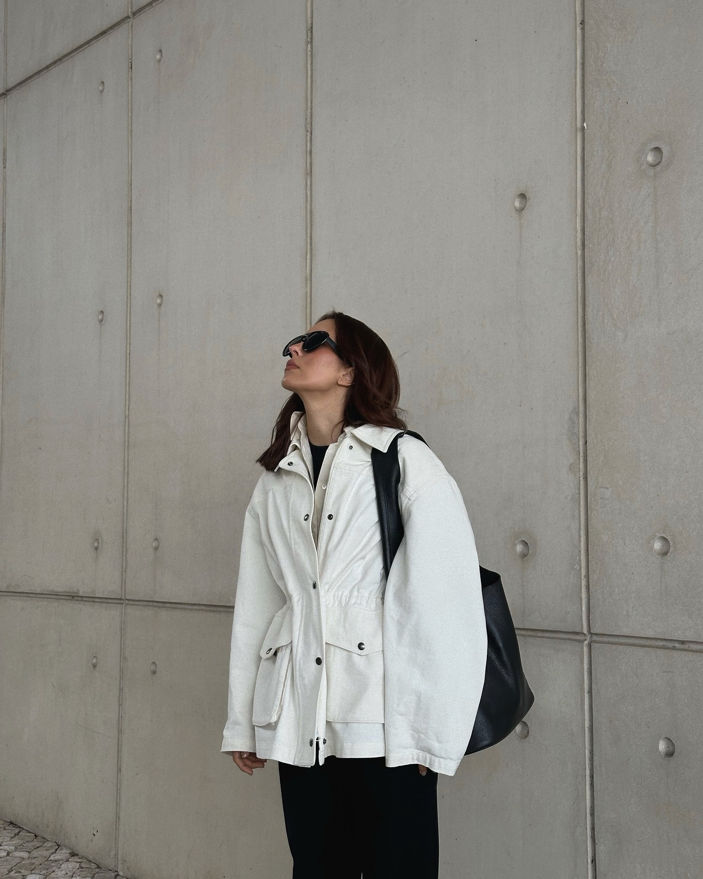 Woman wearing white windbreaker with black pants, standing against wall.