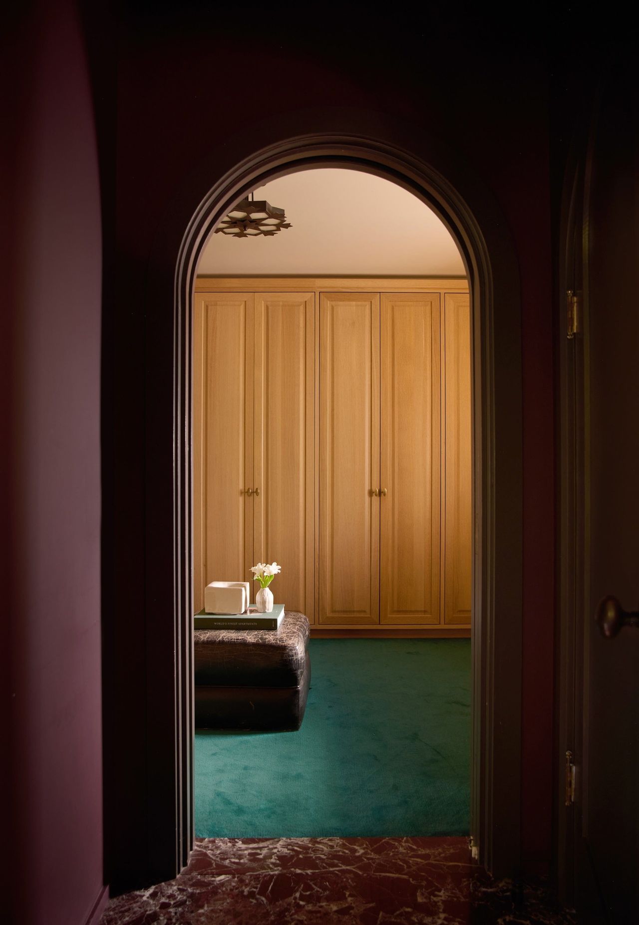 Green carpted dressing room with light wood cabinets photographed through archway with dark purple walls and marble flooring