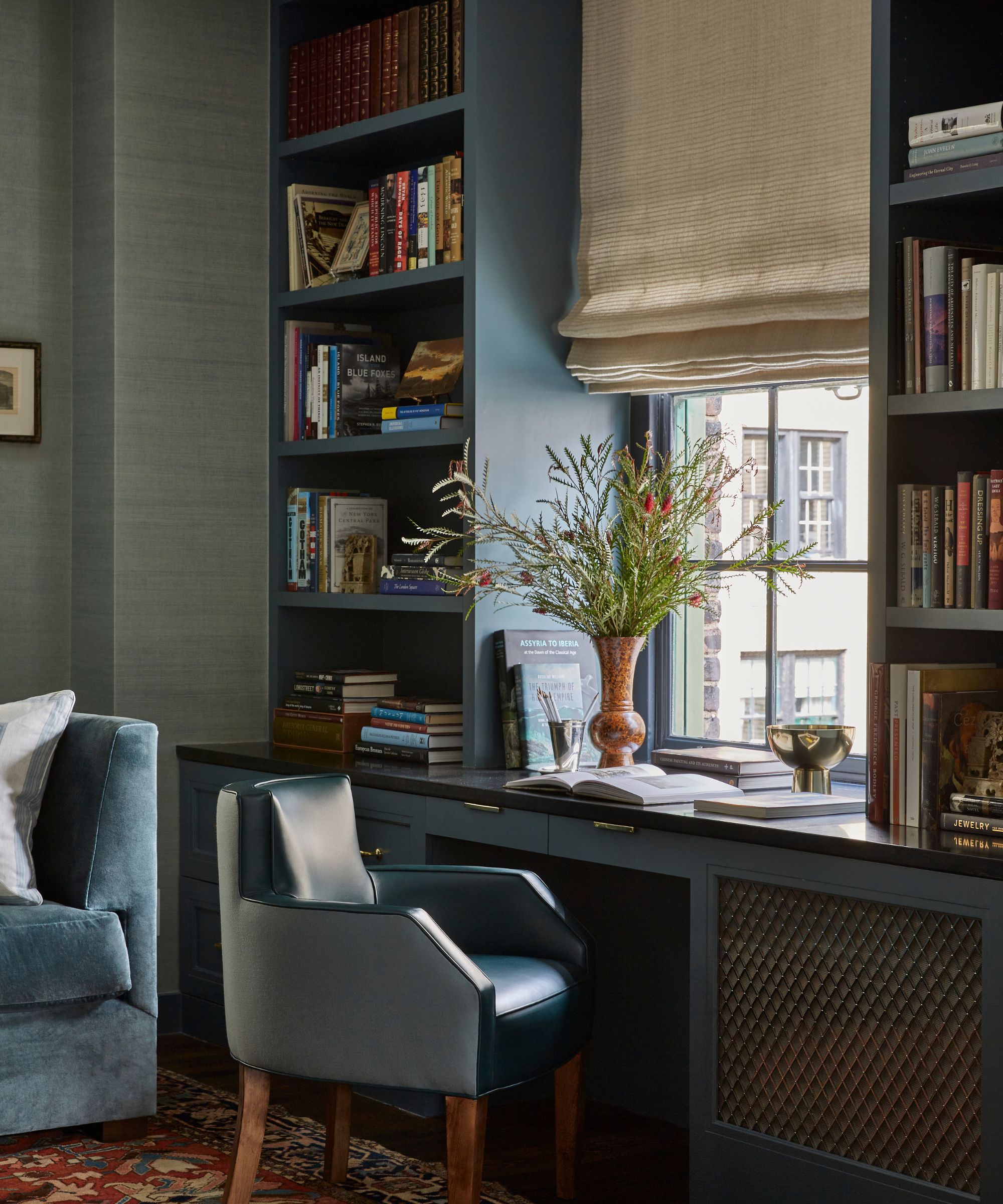 A home office with gray-blue built-ins with a desk area integrated, a cream Roman blind, and a leather armchair.