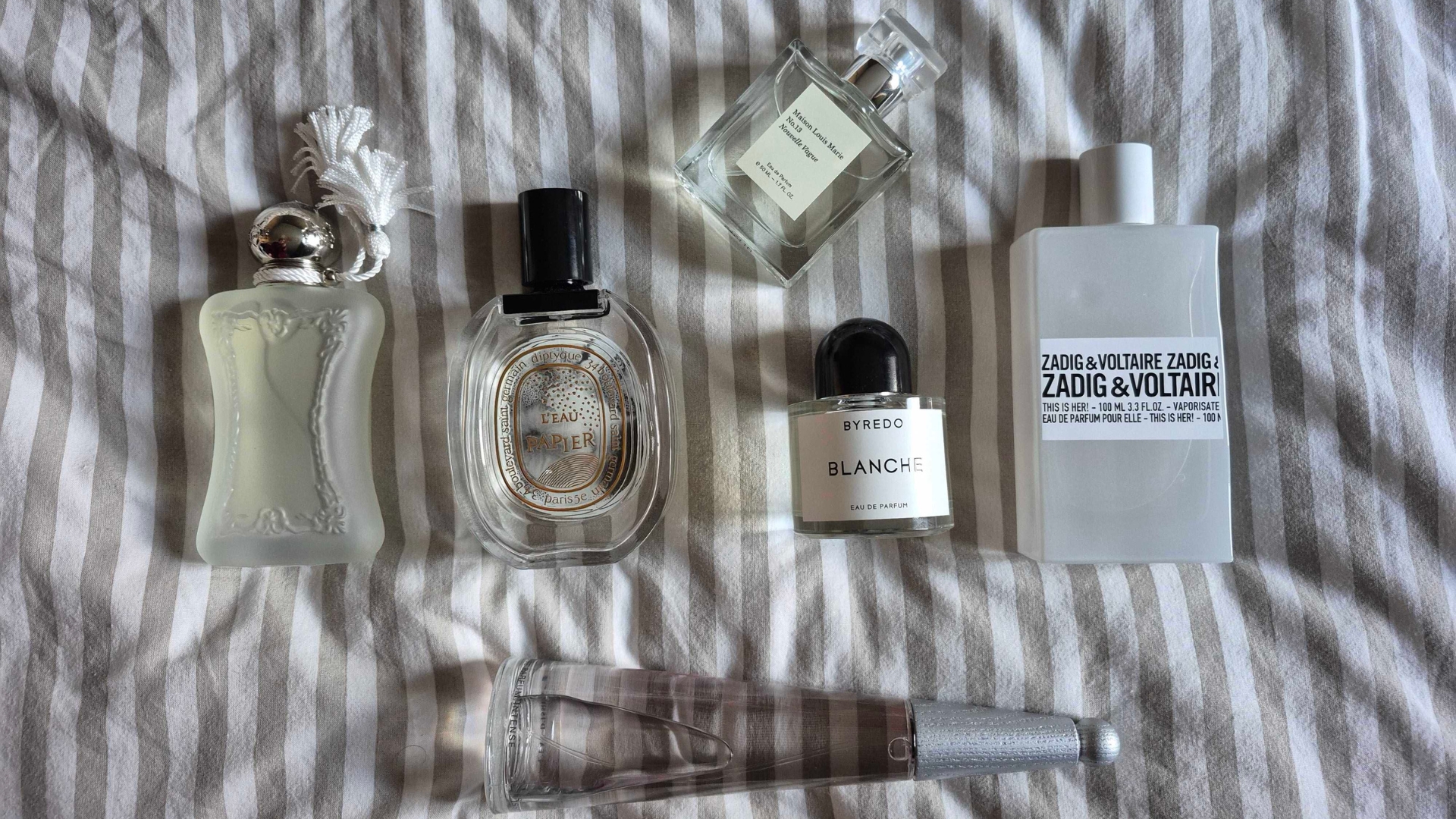 A collection of some of the 'airy perfumes' featured in this roundup, including Parfums de Marly's Valaya, Diptyque's L'eau Papier, Issey Miyake's L'eau D'issey, Byredo Blanche, Zadig &amp;amp; Voltaire This is Her! and Maison Louis Marie No.13 Nouvelle Vague - all laid out and pictured on a white and taupe striped bedsheet background