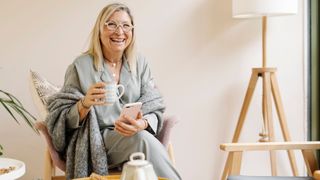 Woman laughing and smiling holding cup of tea and wrapped in blankets at home