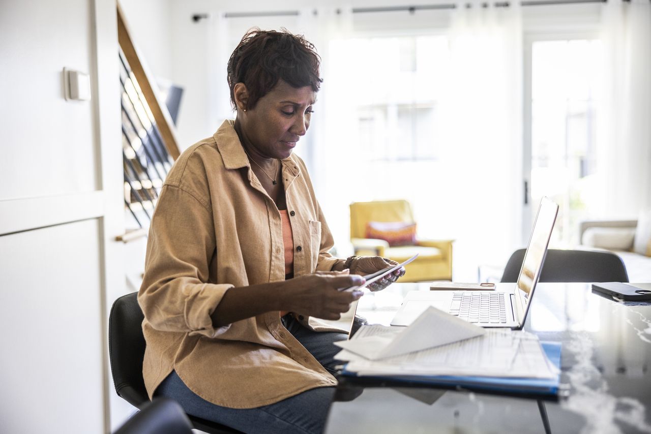 Woman looks at document in her hand as she sits with laptop on kitchen table 