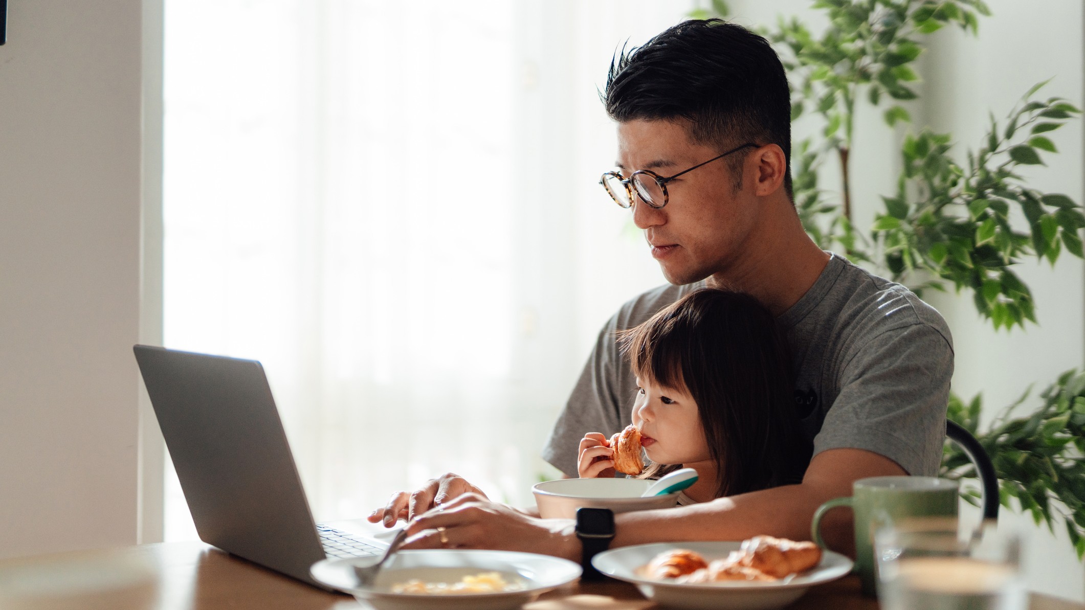 A man uses a laptop at a kitchen table, with his daughter sitting on his lap, eating breakfast. 