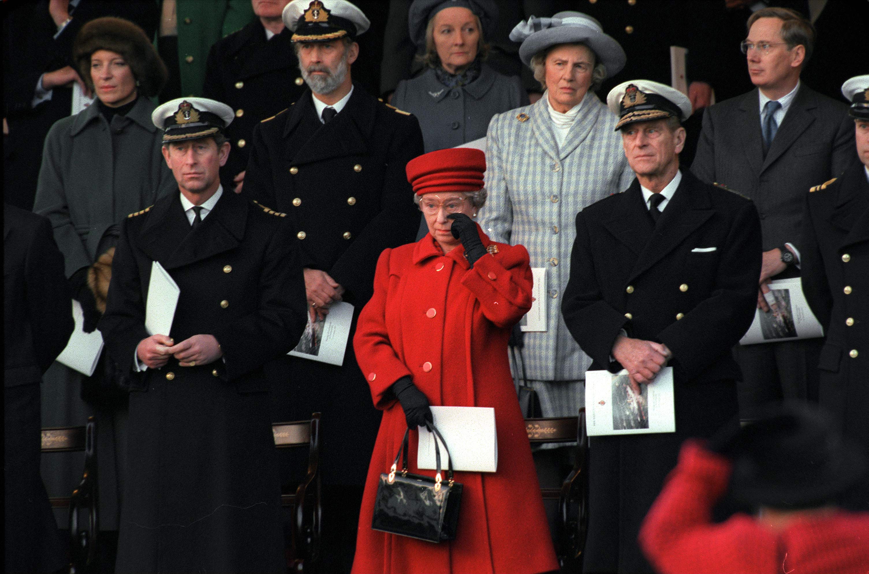 Queen Elizabeth in a red coat wiping her eyes standing next to Prince Philip and Prince Charles