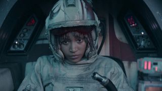 A young woman wearing a well-used pilot helmet and suit is sitting the cockpit of a small space ship.