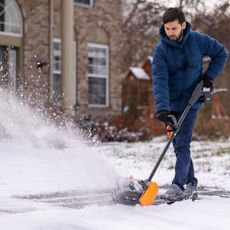 Man wearing blue coat and jeans used Wen cordless show shovel in front of house