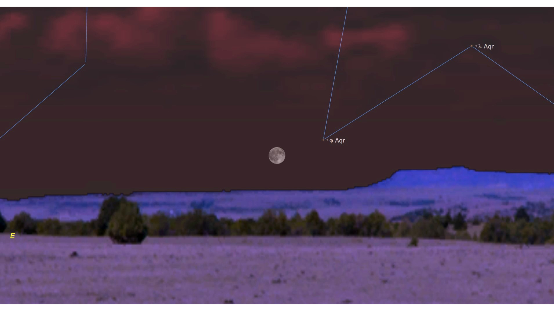 A full moon is seen just above the mountainous horizon far from a rural field in the foreground with a dark red night sky above
