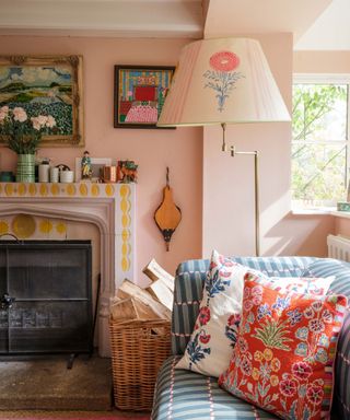 A colorful pink living room with patterned cushions, a tiled fireplace, artwork on the walls, and a shaded floor lamp with floral decor.