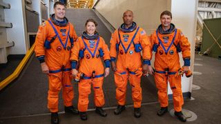 four astronauts stand in a row with orange flight suits on. they are smiling. surrounding them is the deck of a large military boat