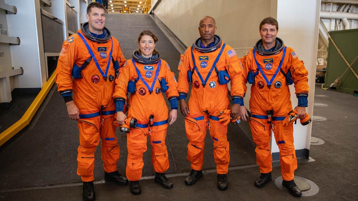 Cuatro astronautas con trajes de vuelo naranjas en una rampa.