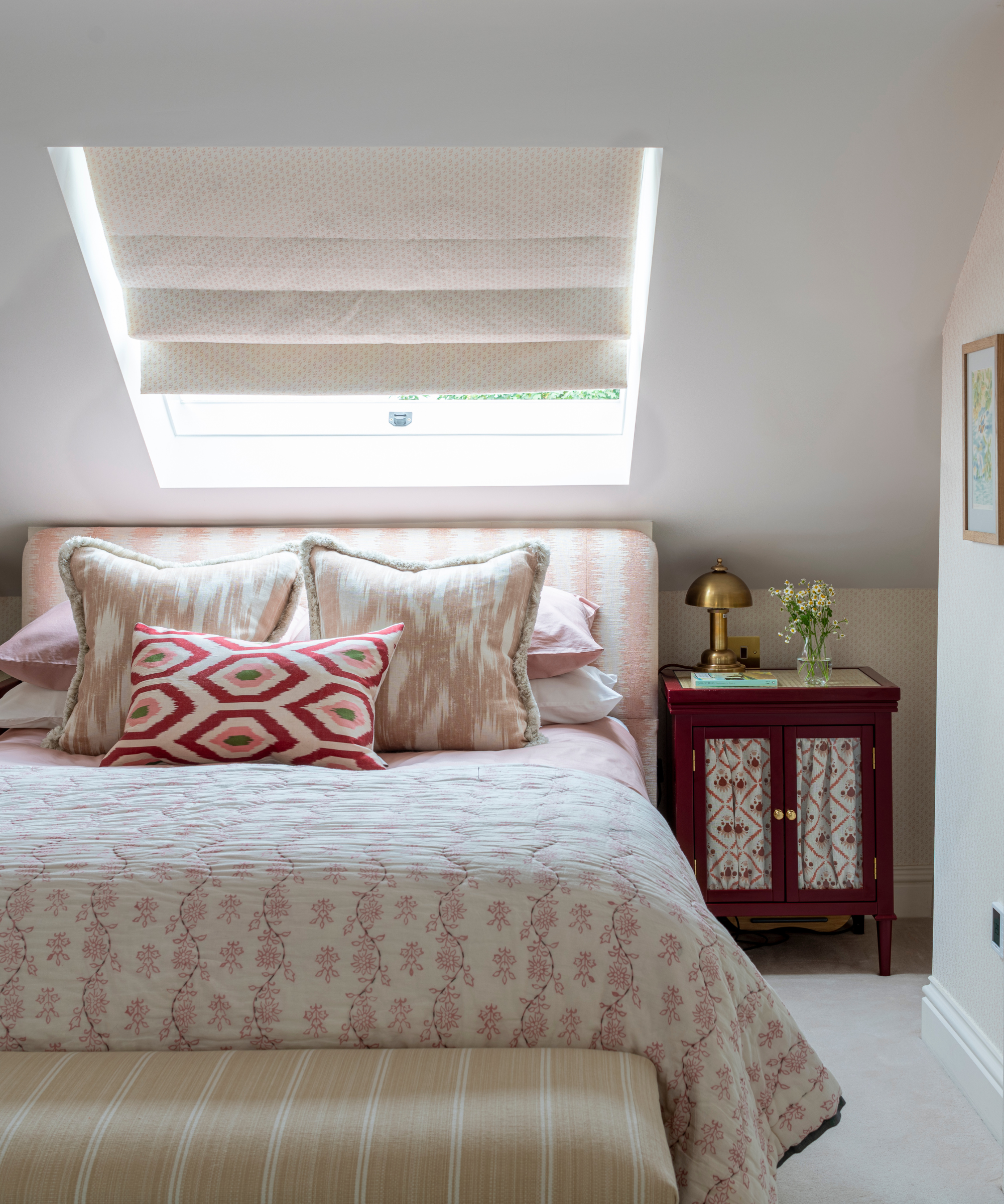 a bedroom with pink floral bedding, an angled ceiling with a window on it, a small side table with a gold lamp, and a bench at the foot of the bed