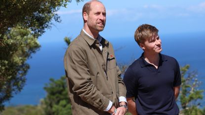 Prince William wearing a tan jacket standing in front of the sea and trees next to Robert Irwin