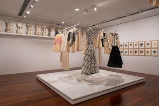 View of the Musée Yves Saint Laurent Paris tribute featuring several fashion pieces hanging on a square rail that is suspended from the ceiling in a room with light coloured walls and wood flooring. There are also half-body mannequins on a shelf and framed art on one wall