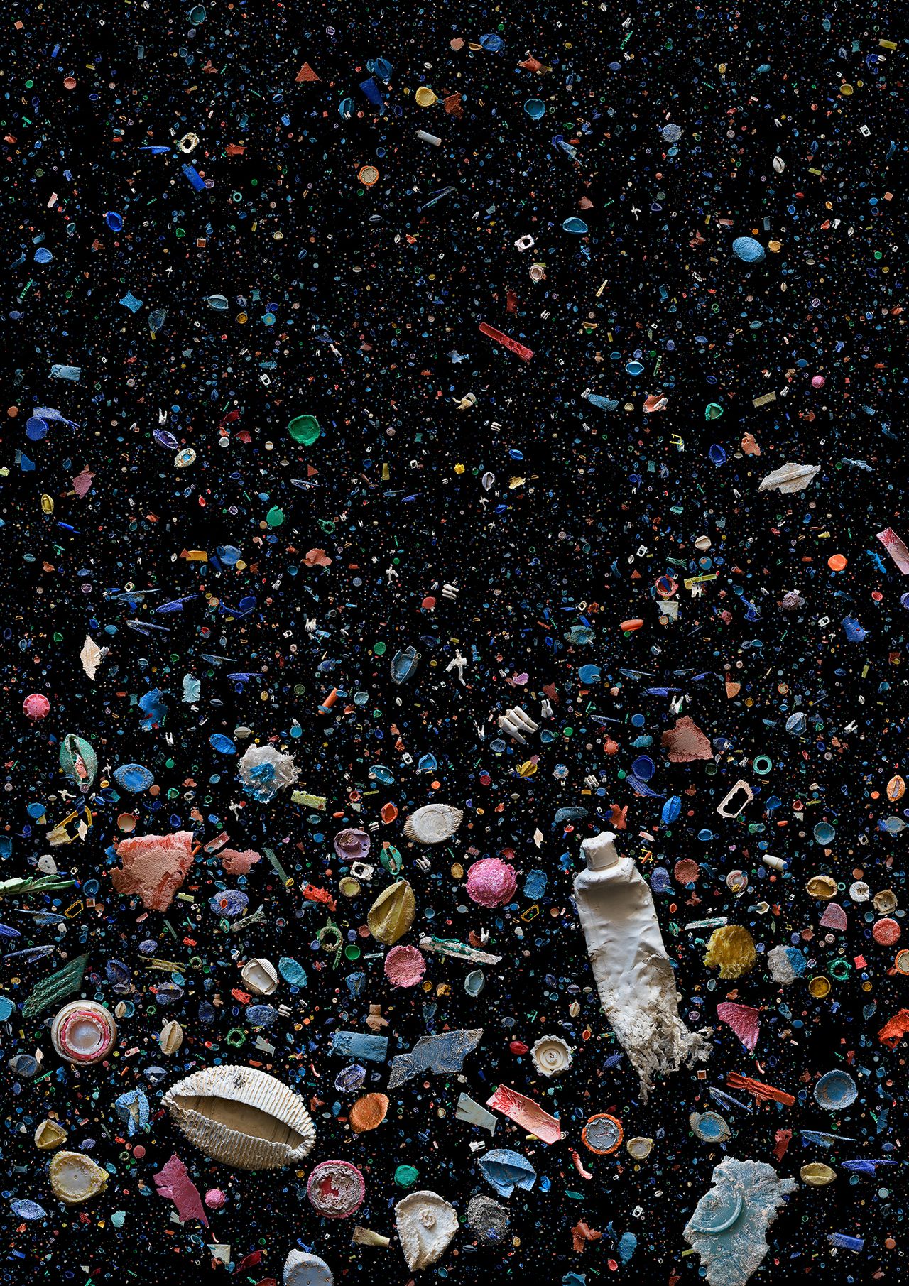 Plastic oceanic debris appears colorfully in sharp contrast against the pitch black of the ocean, items include a toothpaste tube and other recognizable waste