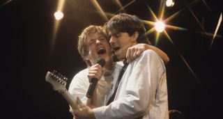 Bryan Adams and Keith Scott share the mic onstage in 1985, with Scott playing an S-style electric guitar.