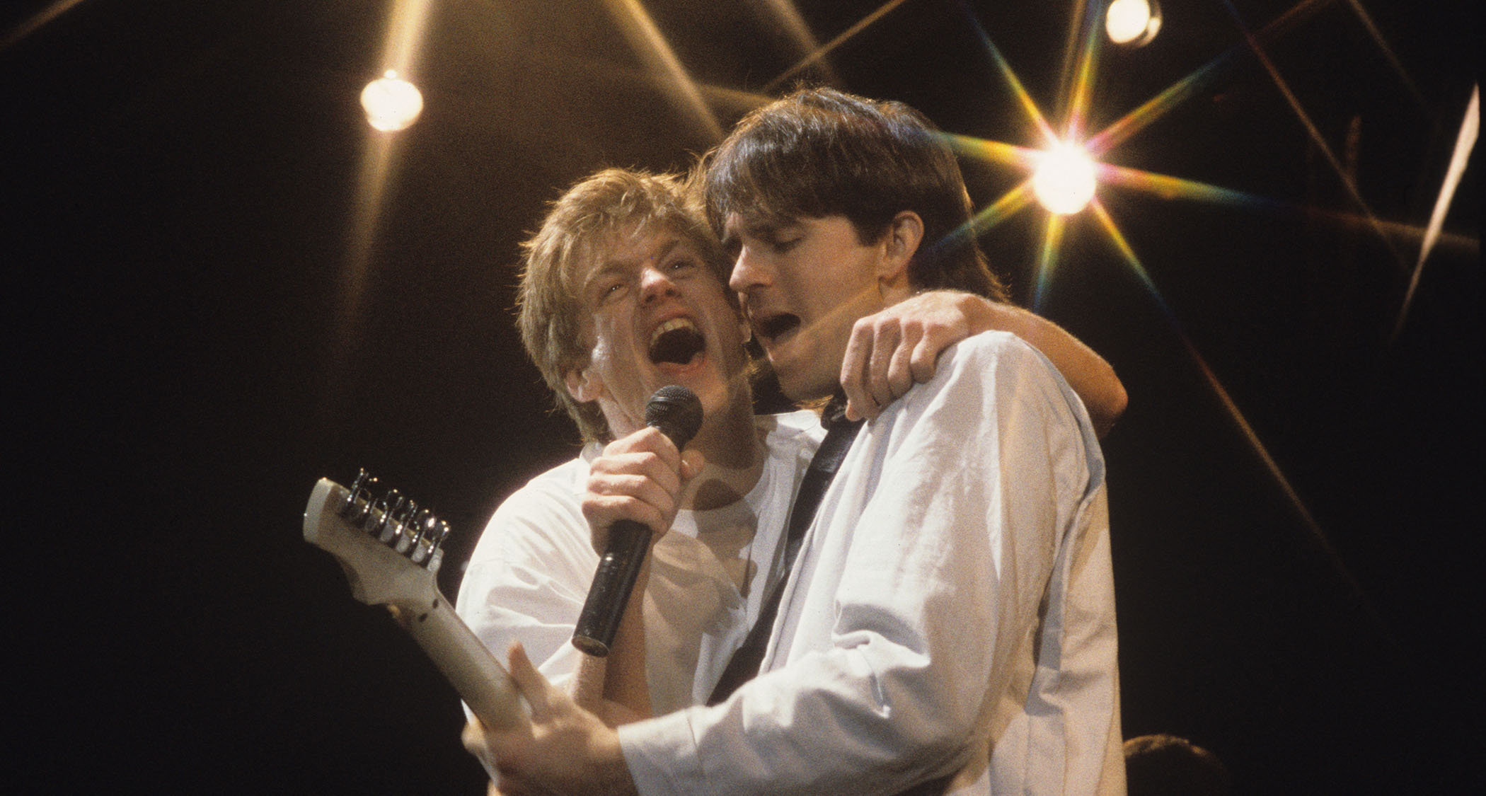 Bryan Adams and Keith Scott share the mic onstage in 1985, with Scott playing an S-style electric guitar.