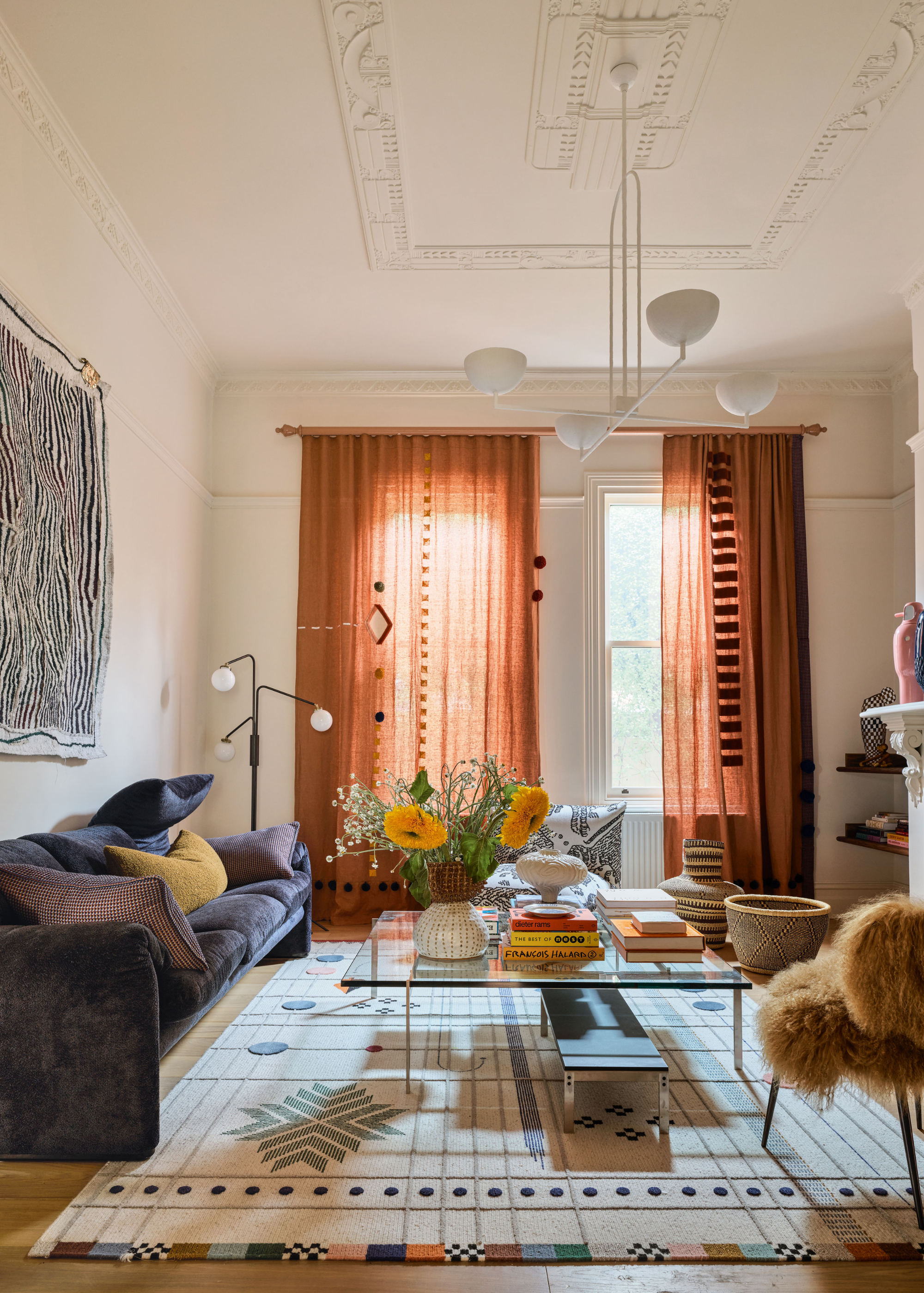 A light, modern, high ceiling living area with tall windows along the far wall, dressed with custom rust coloured curtains, and a white, industrial style chandelier. On the left wall hangs a monochrome textile piece, above a deep, plum coloured sofa. In the centre of the space is a geometric design rug, with a glass coffee table and modern, fur upholstered chair.