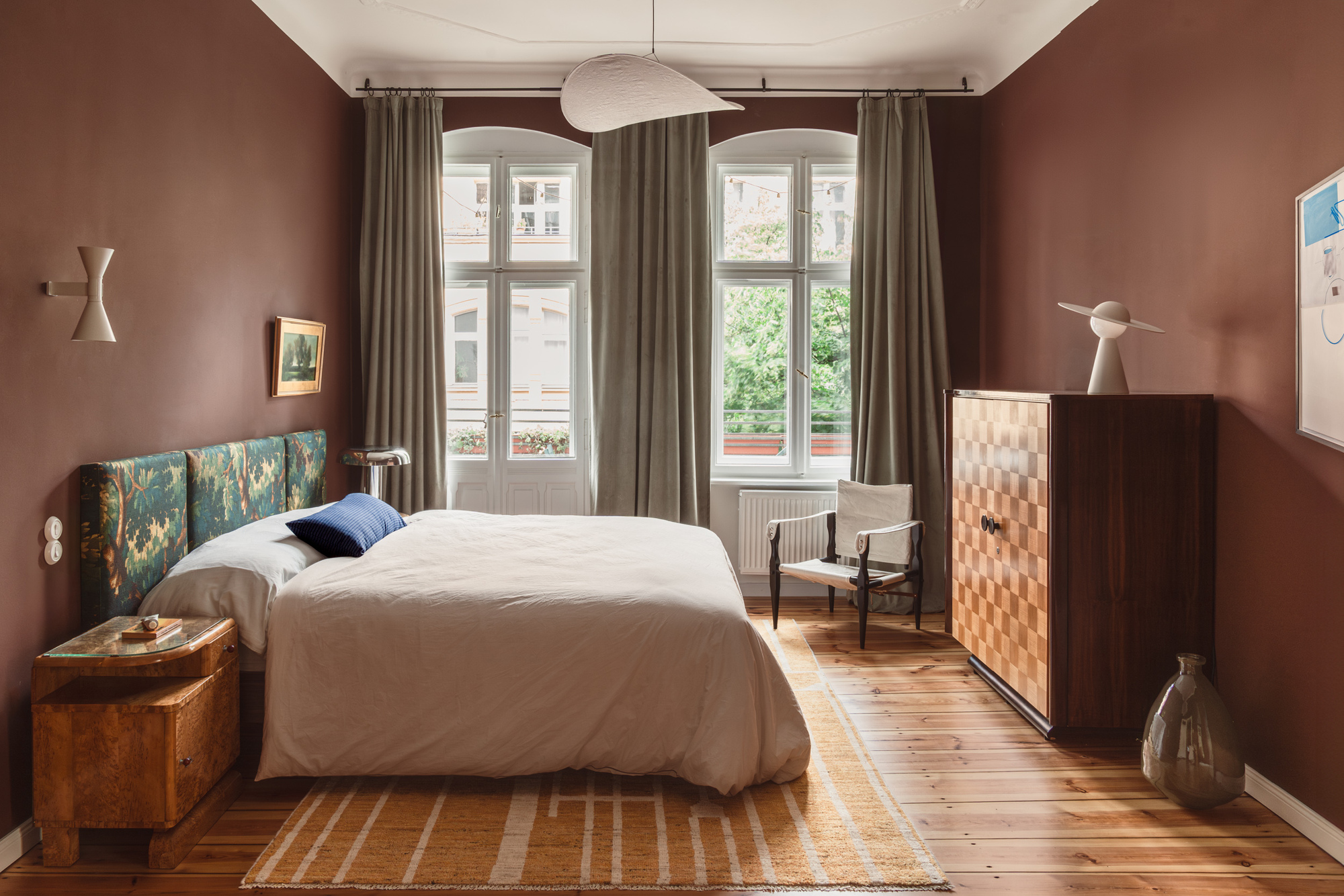 bedroom painted in warm clay color, tapestry headboard, natural linen bedding, timber floors, timber side table, marquetry cabinet with lamp on top and glass vase on the floor beside
