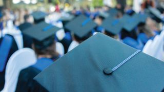A blue graduation cap in focus in the foreground while blue capped graduates are seated in the background