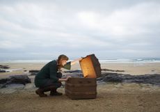 A woman looks inside a glowing treasure chest on a beach.