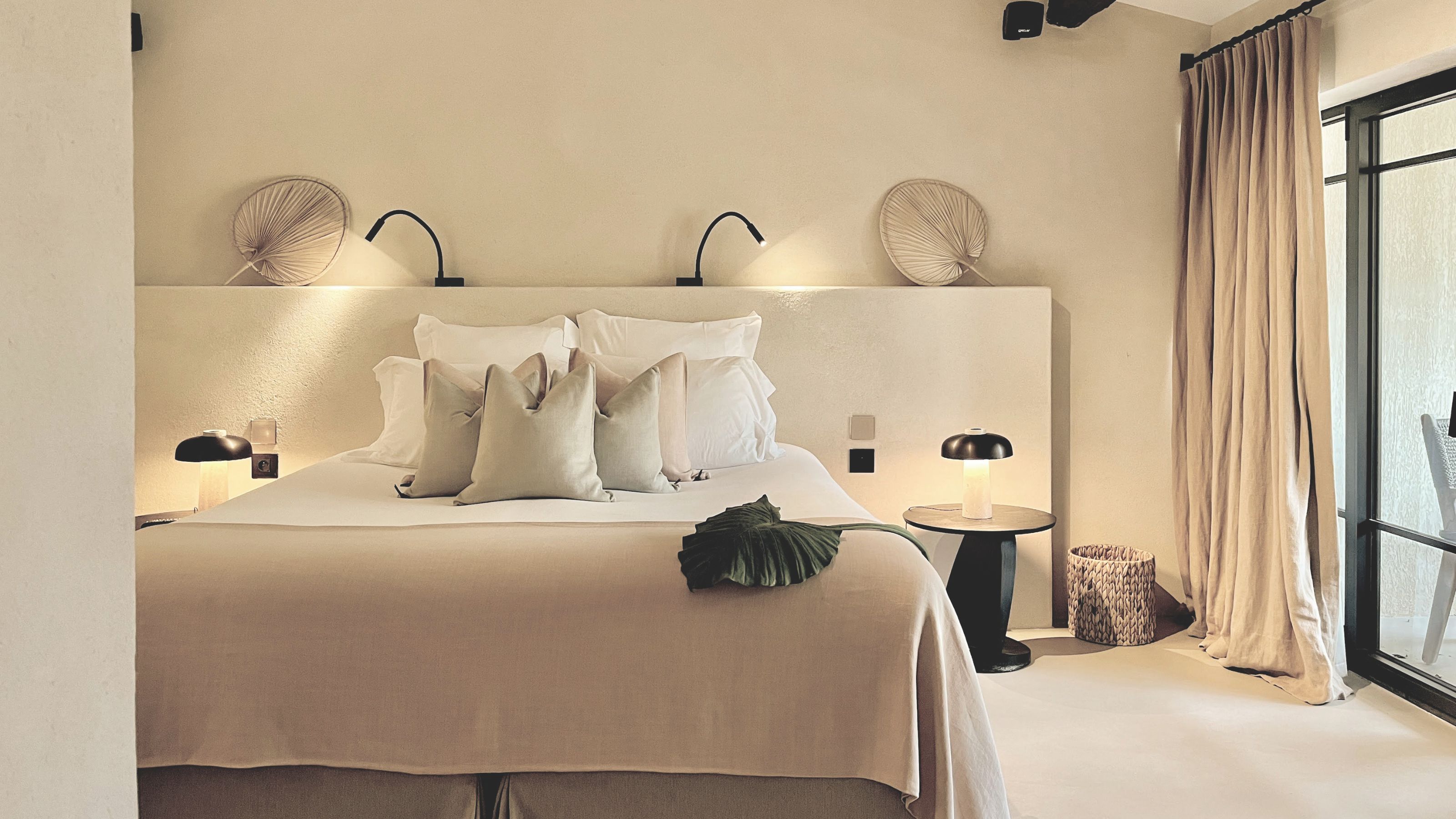A serene, earth-toned suite at Toile Blanche featuring a large bed, symmetrical designer lamps, and floor-to-ceiling linen curtains leading to a private terrace.