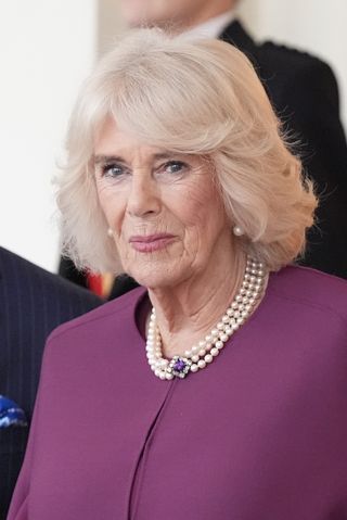 A headshot of Queen Camilla wearing a purple coat and pearl necklace with amethyst clasp