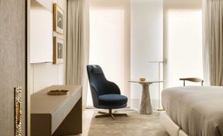 Interior view of a room at The One, Spain featuring light coloured walls, framed wall art, a desk, a dark blue and grey swivel chair, a round table with a white marble effect cone shaped stand, curtains, a rug and a partial view of the bed