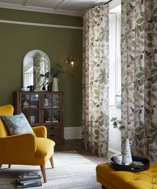 A living room with olive green wall paint decor, mustard yellow velvet upholstered armchair, mustard yellow velvet chesterfield-style coffee table ottoman and curtain window treatments with botanical motif