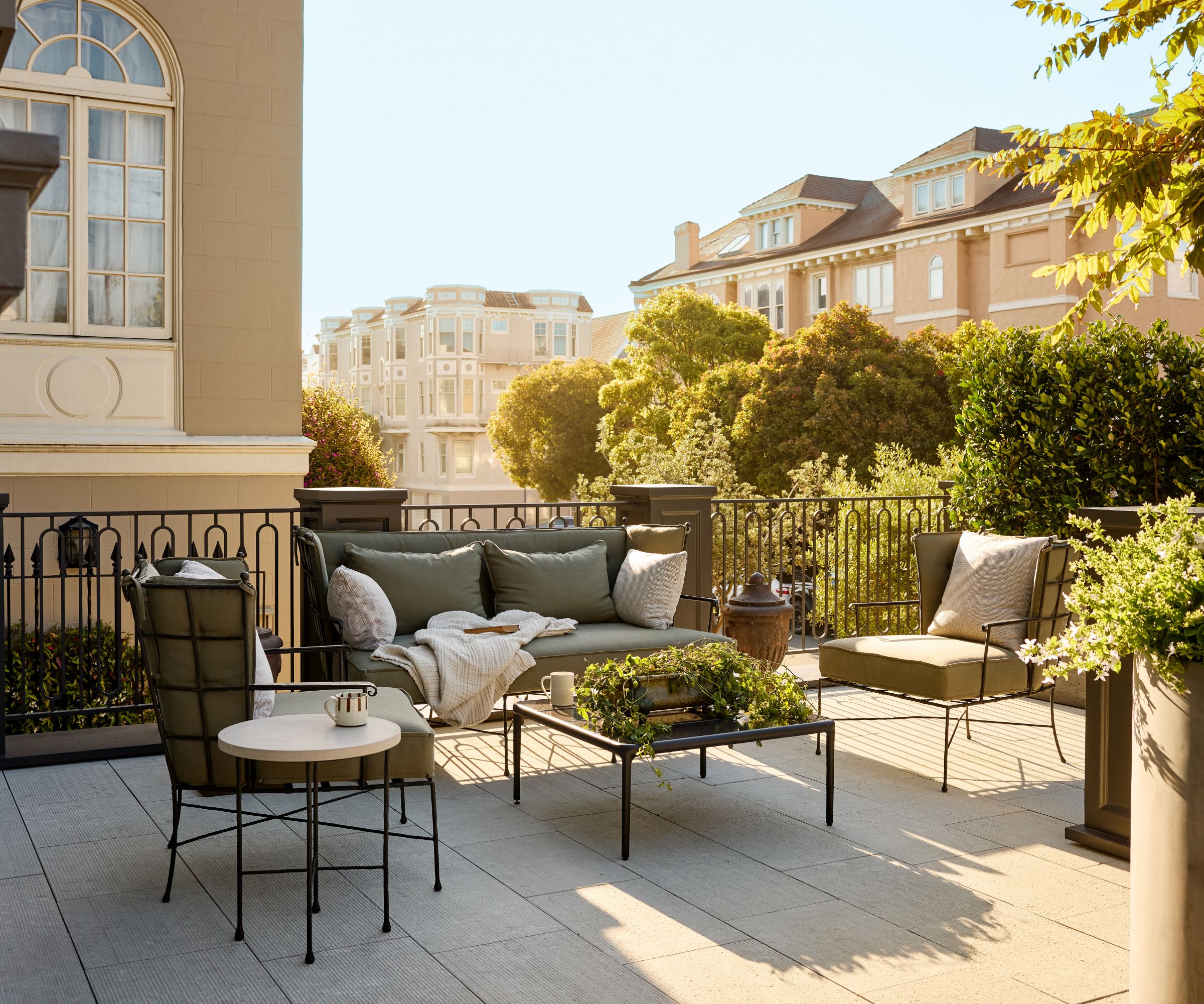 Large terrace with sofa and armchair seating, metal coffee table covered with plant in centre, greenery and buildings in background