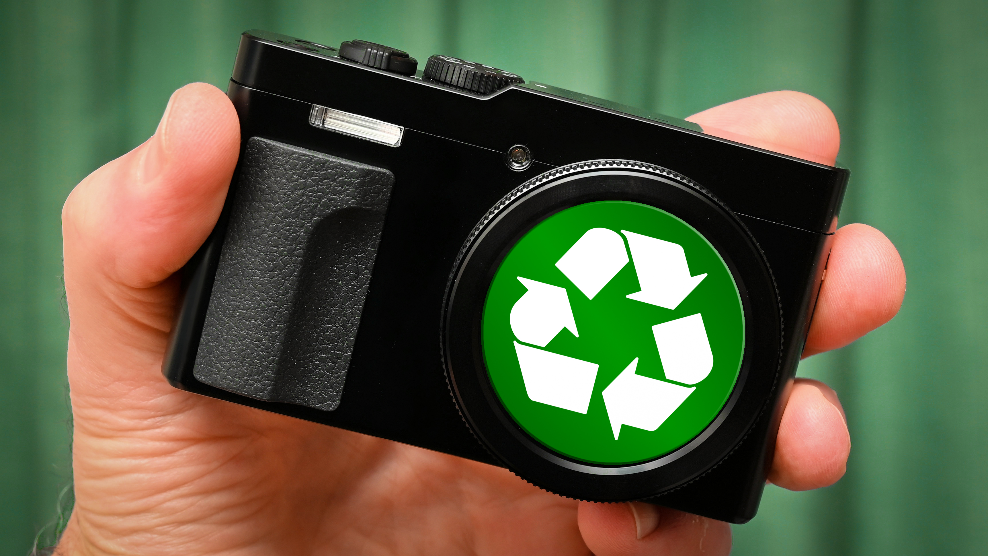 A compact camera with a "recycle" logo on the lens cap, being held by hand