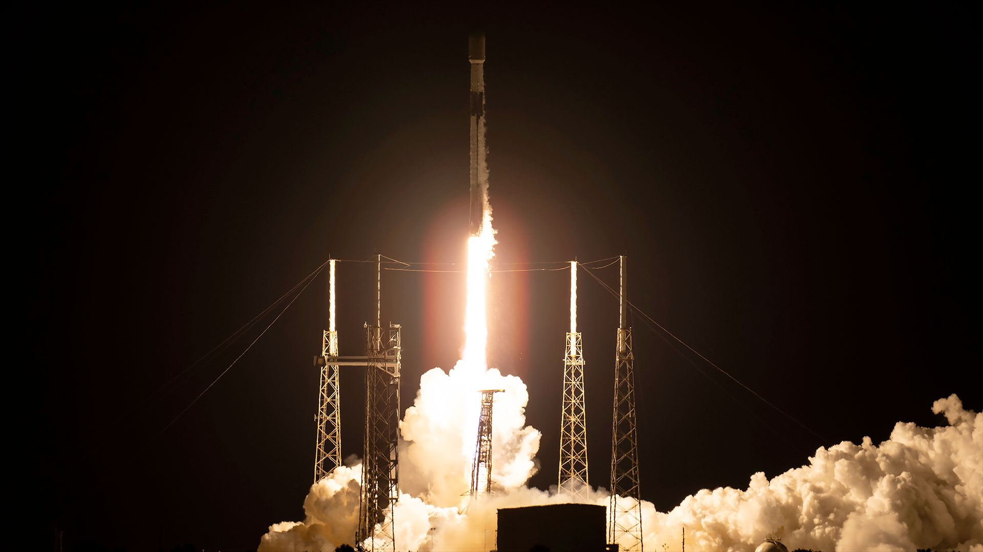 a white and black rocket lifts off into the dark of night, its thrust shining a bright white glow on its launch pad
