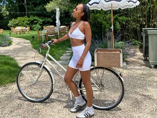 grace wears white sports bra and white biker shorts while holding abike.