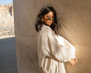 A woman posing in front of a concrete wall wearing round metal sunglasses and a cream-colored balloon-sleeve top