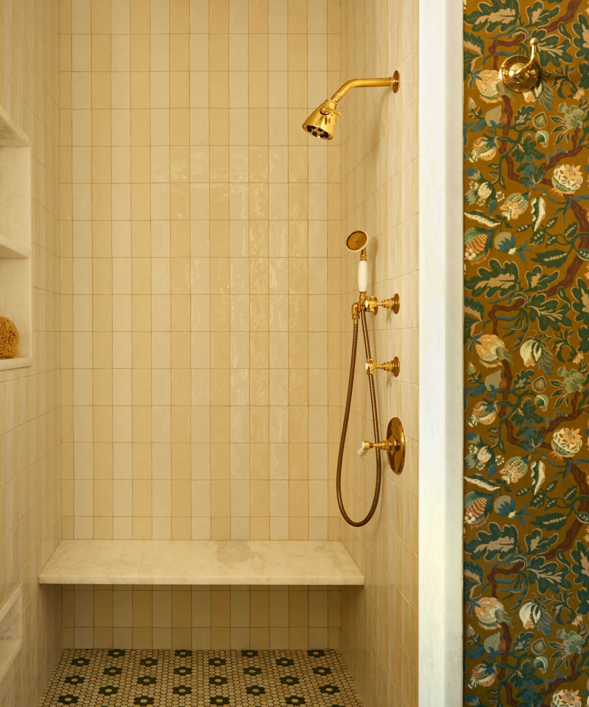 Vintage-style shower with cream vertical tiles, gold hardware, and a marble bench. A glimpse of olive floral wallpaper and penny tile flooring adds a classic, warm touch.