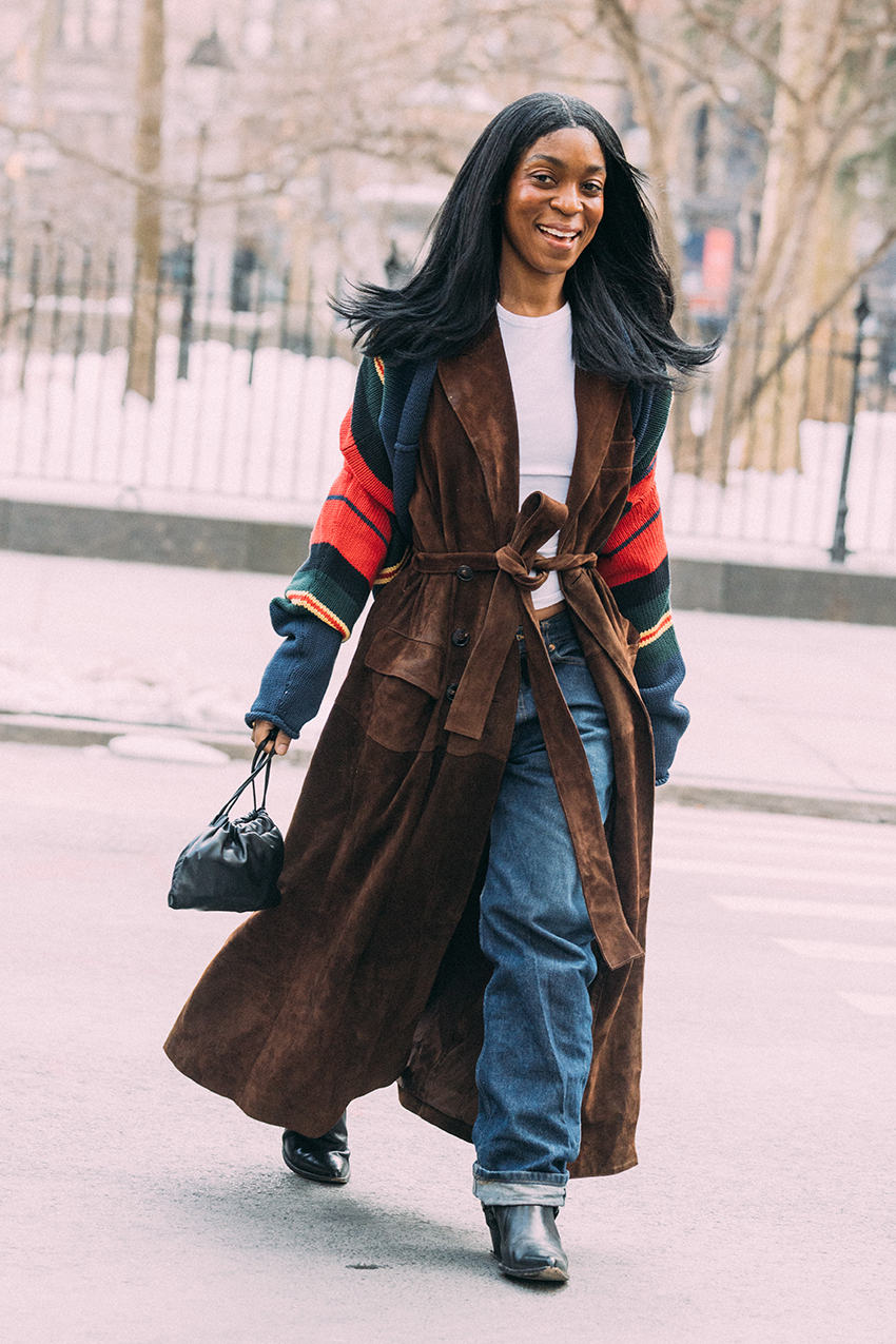 A woman wearing a brown suede coat, white shirt, blue jeans, and black pointed toe shoes