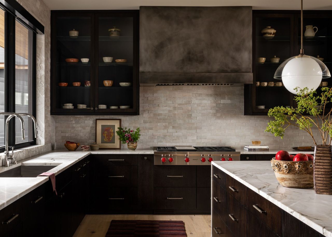 A contemporary kitchen with glass-fronted black cabinets, marble countertops and island, and a gunmetal extractor hood with red knobs on the range cooker