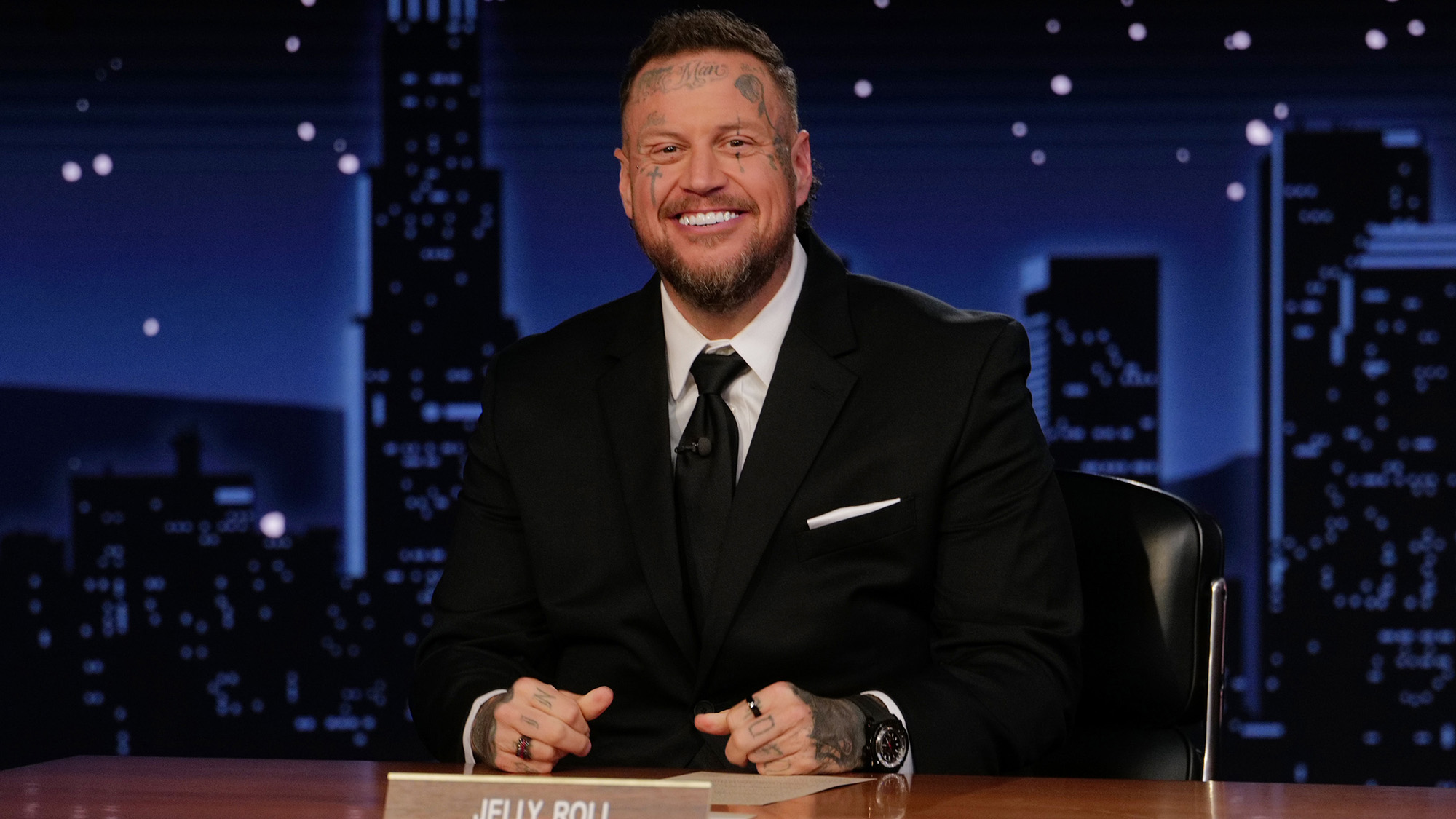 Jelly Roll smiling at the camera while sitting at Jimmy Kimmel's desk on Jimmy Kimmel Live!