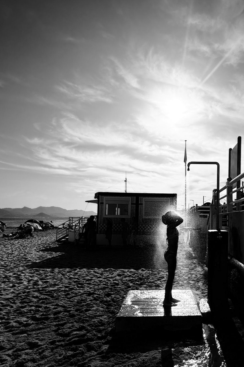 Monochrome image of a young person using an outdoor shower at the beach, silhouetted against a strong backlight