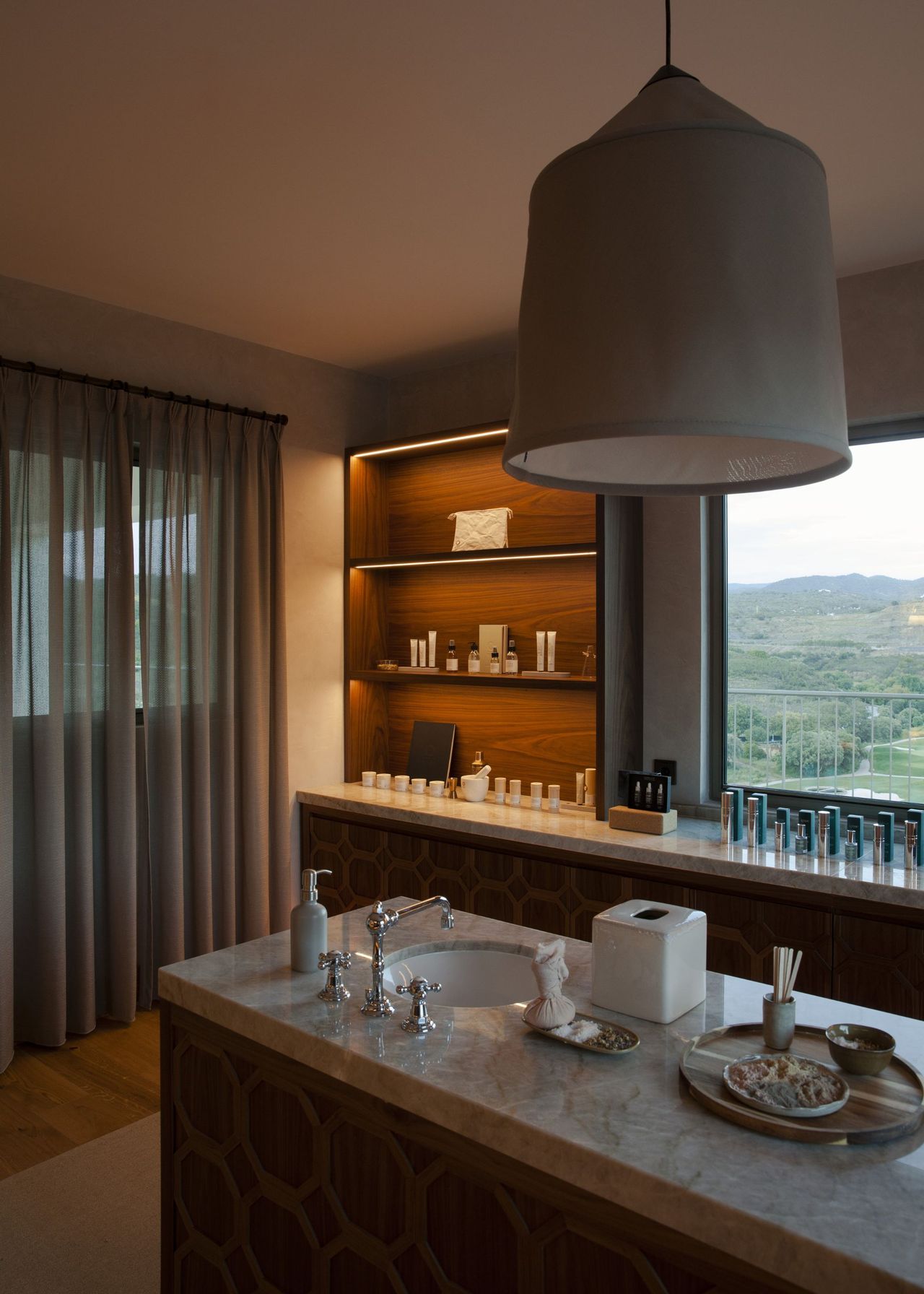 A spa with an island in the middle that had a sink and pendant light hanging above. Behind the island is open shelving with a bunch of creams and treatment oils.
