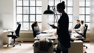 Image of office space with three colleagues sat at workstations in front of a window, and female worker walking across the office with a tablet